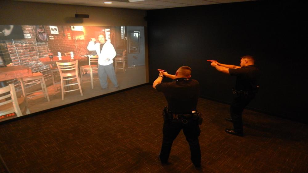 Two officers engage in firearm training simulation, confronting a projected suspect.